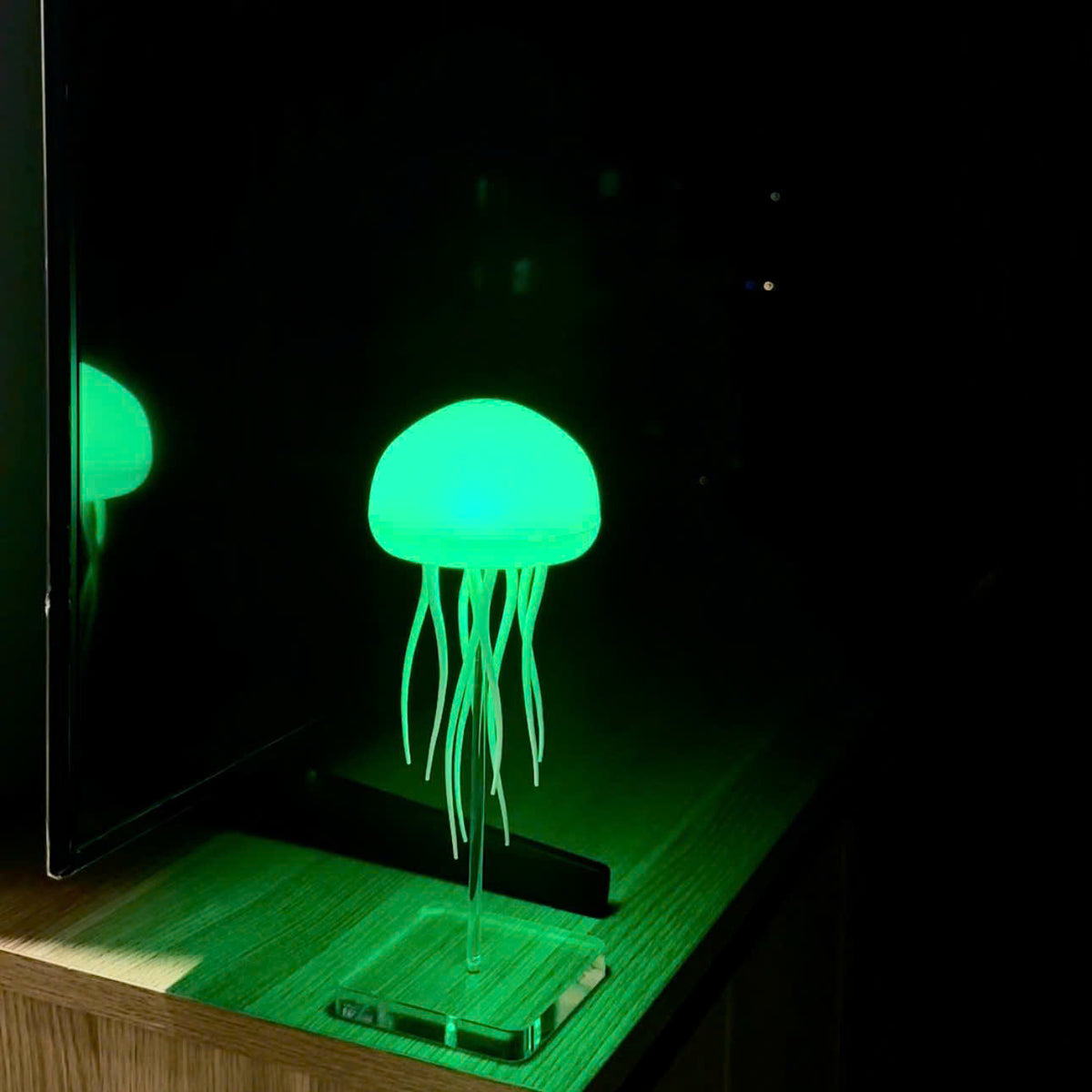 A glowing jellyfish-shaped table lamp with translucent tentacles on a clear acrylic stand, placed on a wooden surface in a dark room with a TV reflection visible in the mirror.