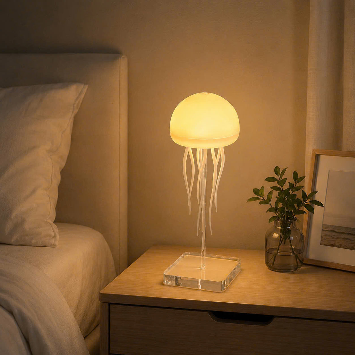 A glowing jellyfish table lamp with a translucent dome and long tentacles sits on a wooden bedside table next to a bed, with a small plant and framed picture nearby.